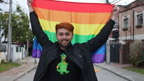 Jesús Ruiz is smiling at the camera and holding up a Pride flag behind him